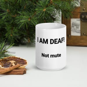 May include: White ceramic mug with the text "I AM DEAF! Not mute" in black. The mug is on a white surface with a dried orange slice and cinnamon sticks.