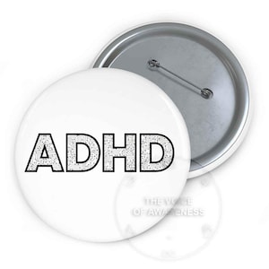 May include: A round pinback button featuring a white background with the black text ADHD, where the letters are filled with a circuit board pattern. The back of the button is metallic silver with a pin mechanism for attachment to clothing.