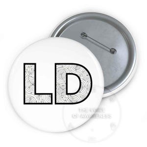 May include: A round pin-back button featuring the black capital letters LD on a white background. The letters are filled with a black and white circular line pattern. The back of the button is silver metal with a pin mechanism for attachment.