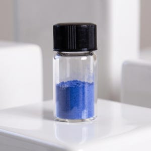 May include: A small, clear glass vial filled with vibrant blue powder, topped with a black, ribbed screw-on cap. The container is cylindrical with a flat base, showcasing the fine, granular texture of the blue pigment stored inside the vessel.