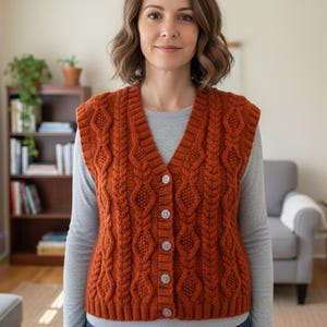 May include: A burnt orange cable-knit vest with a V-neck and button closure. The vest has a textured pattern and is worn over a grey long-sleeved top. The background includes a bookshelf, a grey armchair, and potted plants.
