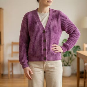 May include: A purple, ribbed-knit cardigan with a V-neck and button closure. The long-sleeved sweater is worn over a light-colored t-shirt and paired with beige pants. The cardigan has brown buttons and a relaxed fit.