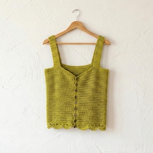 Puede incluir: Un top de ganchillo verde lima con tirantes anchos y una botonadura frontal. El top está colgado de una percha de madera contra una pared blanca. El borde inferior tiene un diseño festoneado.
