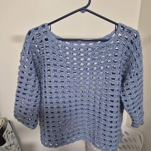 Puede incluir: Un suéter de crochet azul claro con un diseño de tejido abierto. El suéter tiene un cuello redondo y mangas de tres cuartos. Cuelga de una percha negra sobre un fondo neutro.