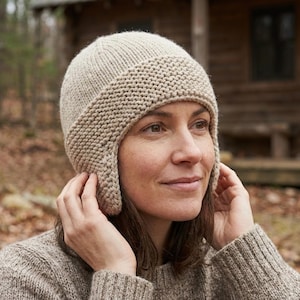 Könnte beinhalten: Eine beige Strickmütze mit Ohrenklappen. Die Mütze ist aus weichem, strukturiertem Garn gefertigt und hat einen gerippten Saum. Die Person auf dem Bild trägt einen passenden Pullover. Der Hintergrund ist eine verschwommene Szene im Freien mit Bäumen und einem Holzgebäude.