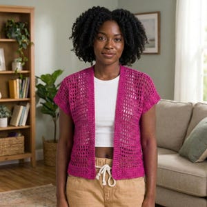 Può includere: Un cardigan all'uncinetto magenta con davanti aperto e maniche corte. Il cardigan è indossato sopra un top bianco e abbinato a pantaloni color cachi. La trama aperta del cardigan è visibile. La donna nella foto ha i capelli scuri e ricci.
