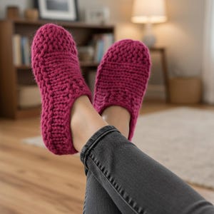 May include: A pair of knitted magenta slippers. The slippers are thick and textured, with a rounded toe and a slip-on design. The person is wearing dark gray jeans. The slippers are resting on a wooden floor.