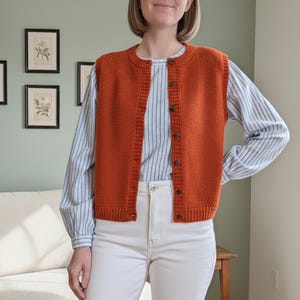 May include: A rust-coloured knitted vest with a button-up front. The vest is worn over a striped shirt and white trousers. The outfit is casual and suitable for everyday wear.