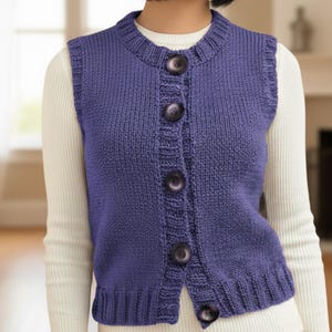 May include: A knitted vest in a periwinkle blue color. The vest has a round neckline and is buttoned down the front with large, dark buttons. The sleeves are sleeveless. The vest is worn over a white long-sleeved shirt.