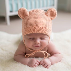 Könnte beinhalten: Ein pfirsichfarbener Babyhut aus Strick mit Bärenohren. Der Hut wird mit einer passenden Schnur unter dem Kinn gebunden. Das Baby liegt auf einem weißen, flauschigen Teppich.