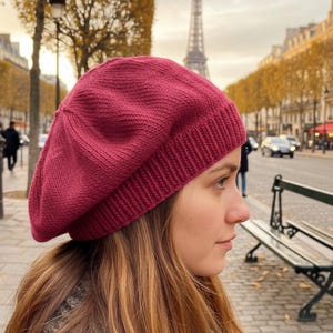 May include: A burgundy knitted beret. The beret is made of a textured knit fabric and is worn on the head. The background shows a city street with the Eiffel Tower in the distance.