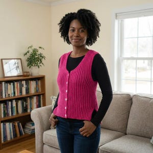 May include: A vibrant pink, button-up knitted vest with a ribbed pattern. The vest is worn over a black long-sleeved top and blue jeans. The person is in a room with a bookcase and a window, creating a cozy atmosphere.