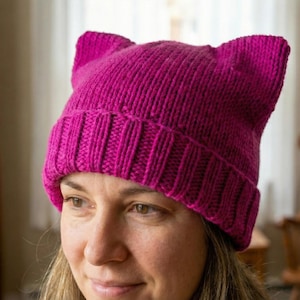 May include: A magenta-colored knitted hat with cat ears. The hat is made of a thick, ribbed knit material. The hat is worn by a person.