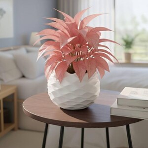 Puede incluir: Una planta en maceta con hojas rosas en un jarrón blanco con diseño geométrico sobre una mesa marrón oscuro. La mesa tiene patas negras. El fondo incluye una cama, una ventana y una pared azul claro.