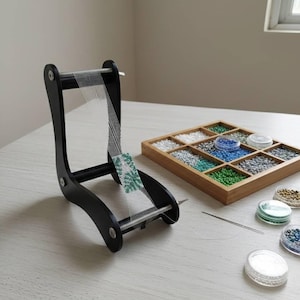 以下が含まれることがあります： 黒い木製のビーズ織り機と部分的に織られたビーズプロジェクト。さまざまな色のビーズが入った木製のトレイと、小さなビーズの容器もテーブルにあります。針も存在します。
