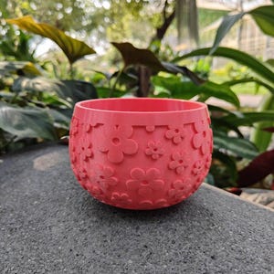 May include: A coral-colored, round planter with a floral pattern. The planter is made of a solid material and features a series of embossed flowers around its exterior. The planter is sitting on a gray surface, with a blurred green background of foliage.