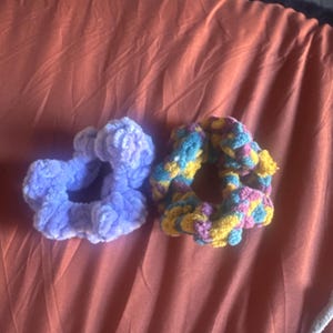 May include: Two handmade scrunchies. One is a solid light purple color, and the other is multicolored with yellow, blue, pink, and purple. Both are made of a soft, plush material.
