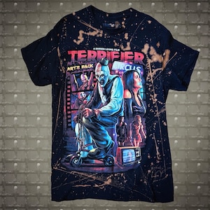 May include: A black short-sleeve t-shirt with a bleach-splattered effect featuring a graphic print from the film Terrifier. The design includes the text A DAMIEN LEONE FILM, TERRIFIER, ART'S BACK, and CIRCUS, depicting a clown on a tricycle.
