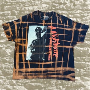 May include: A black short-sleeve t-shirt with a bleach-dyed grid pattern. The front features a black and white graphic of a character in a fedora and the red text A Nightmare on Elm Street. The shirt has a crew neckline and a standard fit.