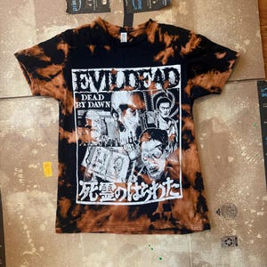 May include: Black t-shirt with a tie-dye effect in shades of brown and orange. The front features a graphic from the movie "Evil Dead: Dead by Dawn" with a skull and other characters. Japanese text is also present.