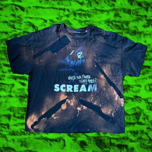 May include: A black short-sleeve t-shirt featuring a bleach-dyed effect with silhouettes of knives. The graphic depicts the Ghostface character from the movie Scream, with the text "What's your favorite scary movie?" and the word "SCREAM" below.