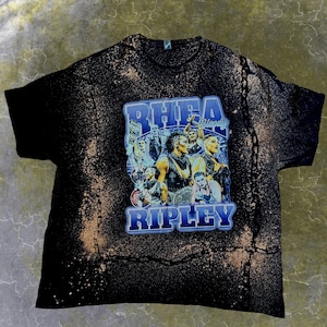 May include: A black short-sleeve t-shirt with a bleached, mottled tan pattern. The centre features a graphic print of professional wrestler Rhea Ripley in various poses with the text RHEA RIPLEY and Bloody in blue, bold, block lettering.