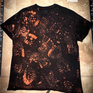 May include: Black t-shirt with a unique design featuring orange-colored shoe print patterns and splatters. The shirt has a crew neck and short sleeves, offering a casual style.
