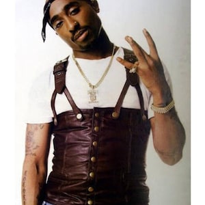 May include: A portrait of Tupac Shakur wearing a white t-shirt, a brown leather vest with gold buttons, and a black bandana. He is also wearing a gold chain and bracelet. The text "TUPAC SHAKUR" is at the top of the image.