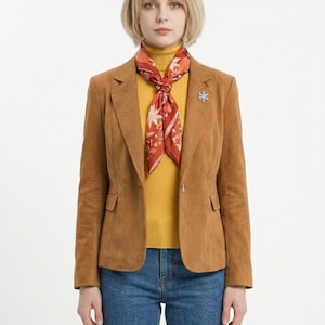 Könnte beinhalten: Ein beigefarbener Blazer aus Wildleder über einem gerippten Rollkragenpullover in Senfgelb. Ein rostfarbenes Halstuch mit weißem Blumenmuster ist um den Hals gebunden. Eine kleine silberne Schneeflocken-Brosche ist am linken Revers befestigt.