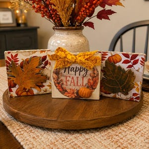 May include: Three square wooden blocks featuring autumn-themed designs. The left block has a large orange leaf, the center block displays a wreath with the text "Happy FALL" and a yellow ribbon, and the right block features a dark green leaf.