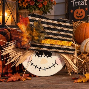 May include: A decorative scarecrow face ornament featuring a round white wooden base with a painted black stitched mouth, closed eyes, and pink cheeks. It wears a black and tan striped burlap hat adorned with a yellow ribbon and a glittery maple leaf.