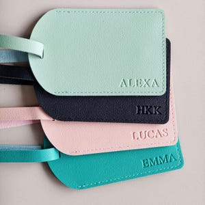 May include: A stack of four rectangular luggage tags with rounded corners, each featuring a matching colored strap. The tags are colored mint green, black, light pink, and teal, with the names ALEXA, HKK, LUCAS, and EMMA embossed on each.
