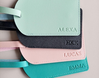 May include: A set of four rectangular luggage tags with rounded corners, featuring a textured finish and matching straps. The tags are coloured mint green with the name ALEXA, black with HKK, light pink with LUCAS, and teal with EMMA embossed.