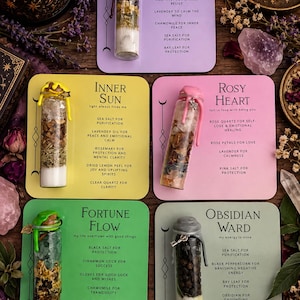 Op de afbeelding: Vijf glazen flesjes gevuld met laagjes kruiden, zouten en kristallen, bevestigd op gekleurde kaarten. De kaarten zijn gelabeld met Calm Within, Inner Sun, Rosy Heart, Fortune Flow en Obsidian Ward, met ingrediënten zoals amethist en lavendel.