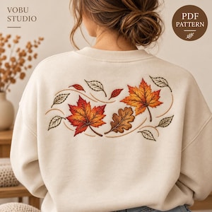 May include: A cream-colored crewneck sweatshirt featuring an embroidered design on the back. The embroidery depicts a collection of autumn leaves in shades of red, orange, and brown, accented by smaller outlined leaves and curved lines.