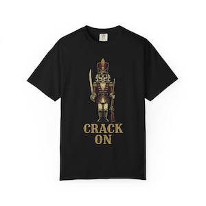 Może przedstawiać: Czarny t-shirt z krótkim rękawem z grafiką szkieletowego żołnierza dziadka do orzechów w czerwonym mundurze ze złotymi akcentami, trzymającego miecz i karabin. Pod żołnierzem znajduje się napis CRACK ON wykonany przetartą złotą czcionką.