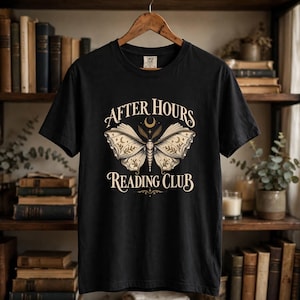 May include: A black short-sleeve t-shirt featuring a graphic of a moth with open wings that resemble book pages. The text "AFTER HOURS READING CLUB" is printed in a cream-colored serif font above and below the moth, with a small crescent moon.
