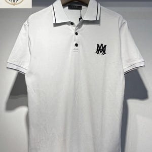 May include: White polo shirt with black trim on the collar and sleeves. Features a three-button placket and a black logo on the chest. The shirt is hanging on a hanger.