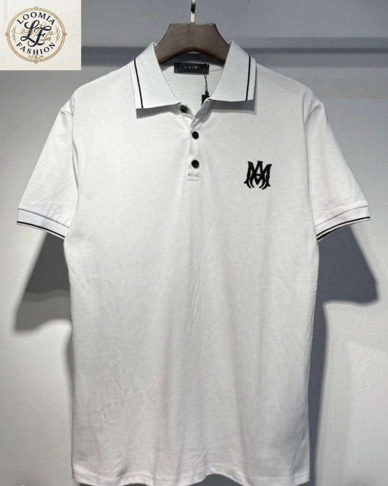 May include: White polo shirt with black trim on the collar and sleeves. Features a three-button placket and a black logo on the chest. The shirt is hanging on a hanger.
