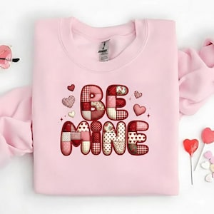 Könnte beinhalten: Ein hellrosa Sweatshirt mit dem Schriftzug "BE MINE" in einem Patchwork-Design. Die Buchstaben sind in roten, weißen und rosa Mustern gehalten. Kleine Herzen und Sterne umgeben den Text, mit herzförmigen Lollis an der Seite.