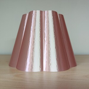 May include: A rose-gold lampshade with a scalloped edge. The lampshade has a glossy finish and a flared, cone-like shape. The design features vertical panels that create a textured appearance. The lampshade is sitting on a light-colored wooden surface.