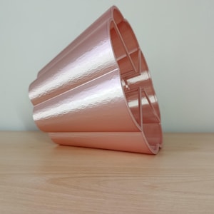 Peut inclure: Un pot de fleurs rose doré métallique à bord festonné. Le pot a une surface texturée et un design ouvert unique. Il repose sur une surface en bois de couleur claire.