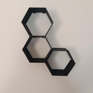 Peut inclure: Une étagère murale noire à trois niveaux en forme d'hexagone. Les étagères sont disposées en cascade, chacune étant reliée à la suivante. Les étagères sont montées sur un mur blanc.