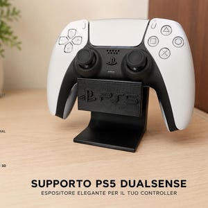 Può includere: Un supporto per controller stampato in 3D di colore nero con il logo PS5 sulla parte anteriore, che sostiene un controller di gioco wireless bianco e nero. Il supporto ha una base rettangolare e un braccio verticale, con il testo PS5 DUALSENSE sotto.