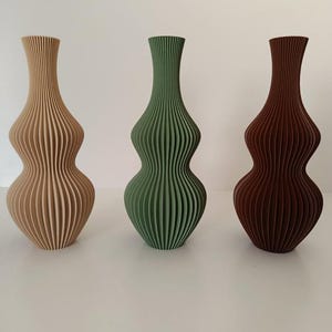 Può includere: Tre vasi decorativi in fila. I vasi sono nelle tonalità del beige, del verde e del marrone. Ogni vaso ha un design a coste unico e una forma curva. I vasi sono vuoti e poggiano su una superficie bianca.