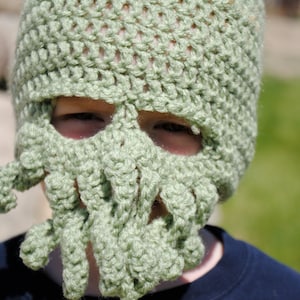 May include: A green crocheted hat shaped like a skull with tentacles. The hat is worn by a person, but only the hat is visible in the image.