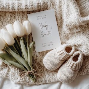 May include: A pair of cream-coloured knitted baby booties beside a card reading "Baby on the Way" with the name "Baby Smith" and the arrival date "Dec 202X". A bouquet of white tulips is tied with twine.
