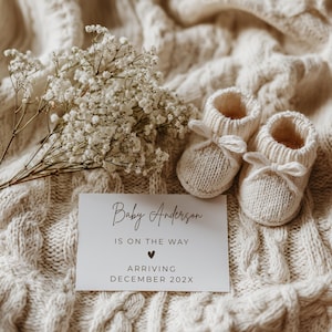 May include: Cream-coloured knitted baby booties with bow detailing, beside a card reading "Baby Anderson is on the way, arriving December 202X." The scene is set on a cream-coloured, textured blanket, with white flowers.
