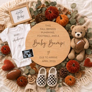 May include: A baby announcement set featuring a white onesie with 'Baby Anderson' text, a round wooden sign reading 'This fall brings pumpkins, football, and a baby bump! Due to arrive April 2026', a crochet teddy bear, mini pumpkins, and baby shoes.