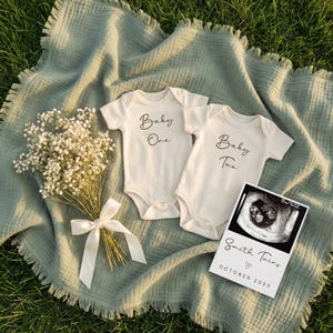 Può includere: Due body da bambino color crema con le scritte "Baby One" e "Baby Two" in corsivo. Un bouquet di fiori bianchi legato con un nastro color crema e una card con ecografia con scritto "Smith Twins" sono presenti nell'immagine.
